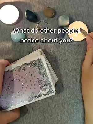 The image features a tarot card reading scene with various elements arranged on a textured gray surface. A deck of tarot cards is held in a person's left hand, its back design visible, featuring a central circular symbol with stars and a silhouette figure. Scattered around are several objects: lit candles, small polished stones in shades of blue, green, and brown, and a silver ring. The text overlay, “What do other people notice about you?” is displayed above the cards.