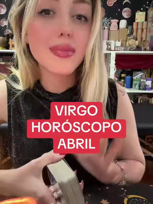 A woman with blonde hair is in the center of the image, looking at the camera with a slight smile. She is holding a deck of cards in her left hand with a red and white text overlay that says, "VIRGO HOROSCOPO ABRIL". The background is a cluttered shelf area with various items.