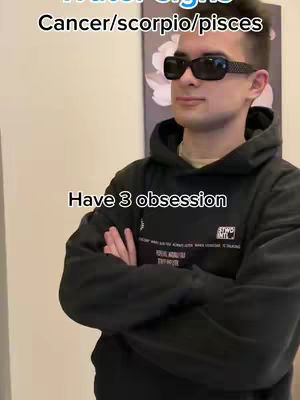 A man wearing sunglasses and a black hoodie stands with his arms crossed, text overlays are placed above and at the bottom of the frame. The background is a blurred room with a piece of artwork. The lighting is natural.