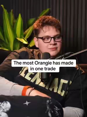 The cover image shows a young man, sitting in what appears to be a podcast or interview setting, with a microphone in front of him. He wears glasses, a black T-shirt with white text, and an orange watch. Behind him is blurred greenery and a dark background. A text overlay reading "The most Orangie has made in one trade" is at the bottom.