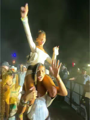 Two young women are at a concert or similar outdoor event. One is being held up on another's shoulders in the center. The woman on top is smiling with her arm up, pointing to the sky. The woman holding her up is also smiling and looking upwards. The crowd around them are blurred from motion.