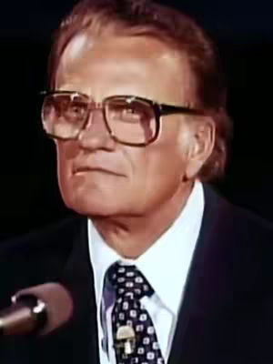 A medium shot of Billy Graham, a man in a dark suit, white collared shirt, and patterned tie. He wears eyeglasses and looks slightly to the side. A microphone is visible near him, suggesting a public speaking setting. The background is darkly lit.