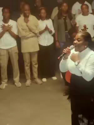A group of people, mostly dressed in white, gathered in a room, with a woman in the front holding a microphone and singing. In the background, there are more people, some with hands raised or clasped; the lighting seems dim with the focus mainly on the woman with the mic.