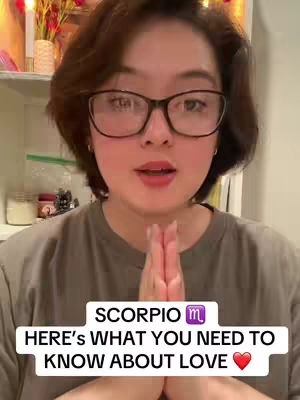 A medium shot features a woman with short brown hair and glasses looking directly at the camera with her hands together in front of her chest, as if in prayer. The background is blurred, hinting at a domestic interior. Text overlays at the bottom of the image state 'SCORPIO m' and 'HERE'S WHAT YOU NEED TO KNOW ABOUT LOVE ❤️.'