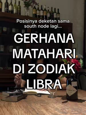 A person is sitting at a table in a room with a bookcase in the background and a bar. The individual is interacting with the camera, a notebook, and a glass of liquid. The focus is on elements related to the zodiac.