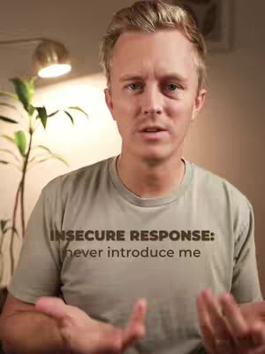A man with light hair and a light olive green t-shirt with the words "INSECURE RESPONSE: never introduce me" written on the front. He has bright blue eyes and is looking at the camera. He is slightly illuminated by light from the bulb of a desk lamp behind him to the upper left of the image.