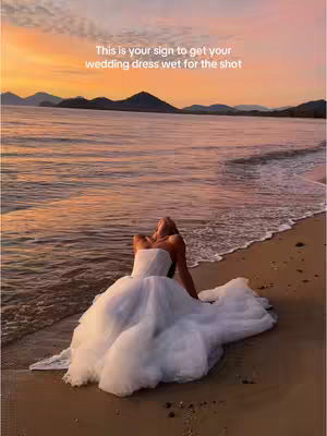 A woman in a white wedding dress is lying on a sandy beach, with the train of her dress spread out. The background features a body of water and an orange and yellow sunset.