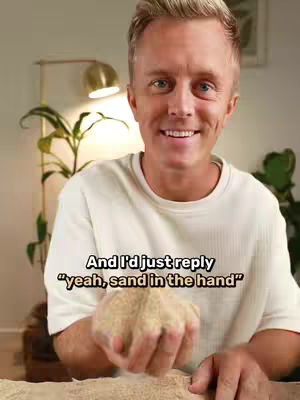 A man with short blond hair and a white shirt is holding a handful of sand, looking at the camera with a smile. The background includes indoor plants and soft lighting.