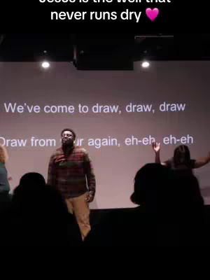 The image shows a live performance scene in a worship setting, with a man in a plaid shirt singing in front of a screen with lyrics displayed. The composition includes figures in the audience with raised hands and the foreground is dark, focusing the viewer's eye on the central figure. The colors are muted with some bright lights.