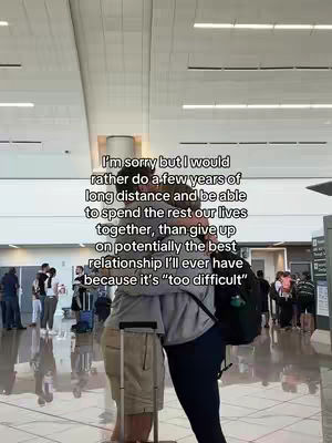 Two people are embracing in an airport, likely a couple. The background shows an airport terminal with signs and other travelers. The image is slightly angled up and is mostly in natural light.