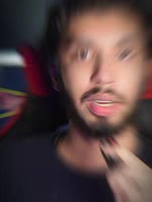 A blurred close-up shot of a man's face. The man has short, dark hair, a beard, and a slightly open mouth. A hand, seemingly the man's own, is visible in the foreground, partially obscuring the neck. The background is out of focus