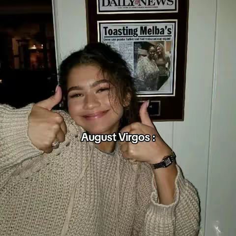 A woman with dark hair smiles and gives two thumbs up. She wears a casual, beige sweater. Behind her, a newspaper clipping is framed on the wall.