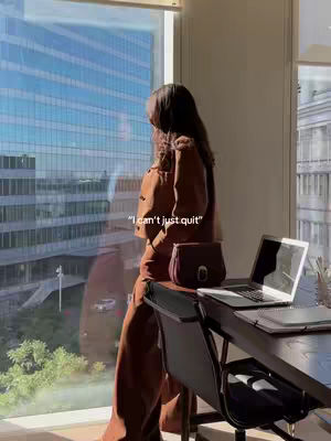 A woman stands in profile near a large window, gazing out. She is dressed in a neutral-toned outfit, including a jacket and pants, which is contrasted by the bright cityscape visible through the window. A laptop and a purse sit on a table beside her. Text overlayed reads, 'I can't just quit'.