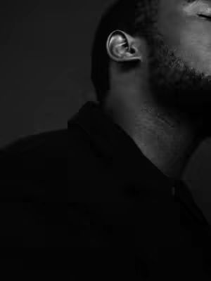 A black and white image shows the side profile of a man. The focus is on his ear and the side of his face, with a dark background. His features are partially visible.