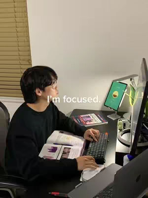 The image shows a person, appearing to be a young man, sitting at a desk. He is wearing glasses and black clothing, seems to be studying. In front of him are books, a keyboard, a computer screen, and a tablet display. The lighting suggests it's night time and the scene has a cozy, focused atmosphere.