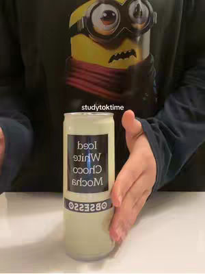 A person is holding a can of iced white chocolate mocha, with the label facing the camera. The background is a black t-shirt with a Minions character on it. The person's right hand is visible, holding the can.