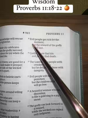The image shows an open book with text from Proverbs. A hand holding a pen points to the text, with an additional text overlay at the top reading 'Wisdom Proverbs 11:18-22.' The color palette is neutral, with the book's pages and the pen's metal prominent.