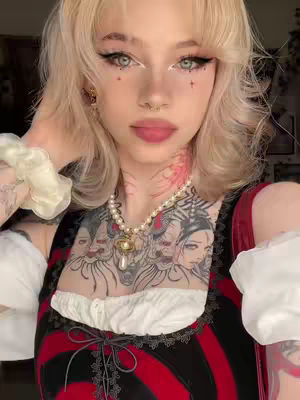 The image showcases a fair-skinned individual with blonde hair and expressive makeup. The person is wearing a red and black corset-style top. A large tattoo is visible across the chest and neck, and a pearl necklace adds an elegant touch. The background is blurred, focusing attention on the person.