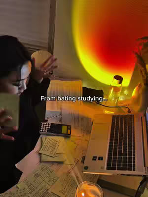 The image captures a dimly lit study space. A young woman is shown from the side, interacting with what appears to be notes and a laptop on a desk littered with papers. The left side of the desk has a calculator and post-it notes. The background features soft lighting with a red and yellow glow, casting a warm, focused ambiance over the scene.