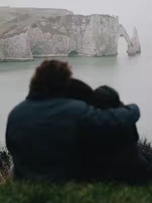 Two people are standing on a grassy cliff overlooking a body of water, with a towering rock formation in the background. The individuals are seen from behind, with their heads touching, partially obscuring the view of the landscape. The sky is overcast, and the overall tone is serene and tranquil.