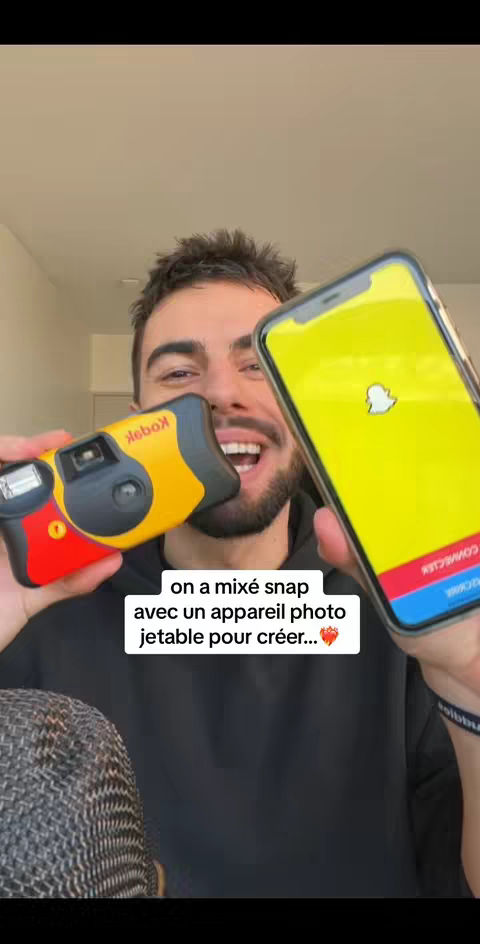 A man holding a yellow and black disposable camera in one hand, and a smartphone displaying the Snapchat interface in the other. The man has a beard and is smiling. The overall composition is a medium shot with a focus on the man and the objects he's holding.