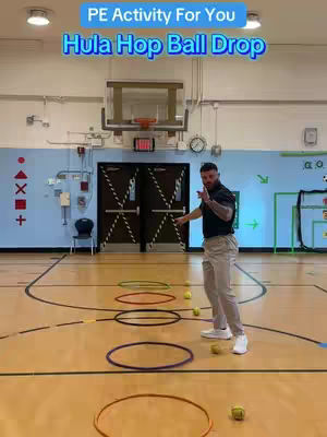 A man in a black shirt and khakis stands on a wooden gym floor, gesturing towards several hula hoops.