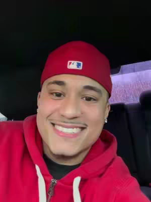 A smiling man in a red baseball cap and hoodie is seated in a vehicle. The person is positioned in the center of the frame, smiling with teeth visible. They have a mustache and earrings. The background seems to be a dark interior, possibly a car.