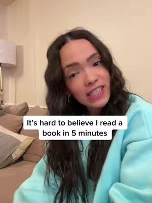 A young woman with long dark curly hair is centered in the frame, looking towards the camera and speaking. She's wearing a light blue sweatshirt, and the background includes a beige wall, a portion of a sofa, and a lamp. Text overlayed on the bottom of the image says 'It's hard to believe I read a book in 5 minutes'.
