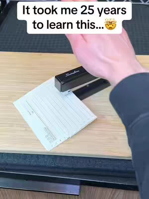 A hand holding a stapler over a piece of paper on a wooden surface. The top of the image has white text over it.