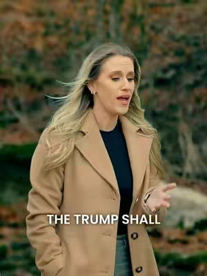 A woman with long blonde hair wearing a light brown coat and a black top is singing outdoors. The background is blurred, indicating a natural setting with trees. White text overlayed at the bottom shows the title of the song.