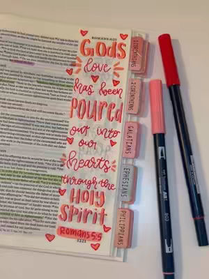 A photo of a Bible open to a page with handwritten text and colorful markers. Handwritten text in orange and pink lettering. The text reads: God's love has been poured out into our hearts through the Holy Spirit Romans 5:5, with multiple little red hearts are drawn throughout the text. A red and pink marker are places on the right side of the bible page. There are bible tabs with different bible book names beside the bible text.