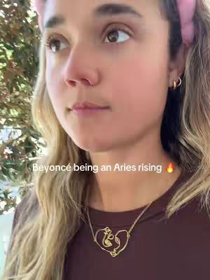 A close-up shot of a young woman with fair skin and light-brown hair looking slightly to the side. She has a pink headband, gold hoop earrings, and a gold necklace in the shape of a heart with what appears to be a hieroglyphic design. The background is out of focus, appearing to be a green foliage. The lighting is soft and natural.