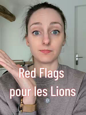 A light-skinned woman with blue eyes, light-brown hair, and pale skin is facing the camera. She has one arm raised in front of her, palm up. Overlayed text in pink and white, "Red Flags pour les Lions", is in front of the chest. The background is a plain white room with a light-colored door and some objects to the left. The woman is wearing a grey sweater and a gold watch with a pendant necklace.