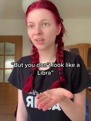 A young woman with bright red hair in pigtail braids is the central focus. She's wearing a black t-shirt and has a slight smile, and her hand is extended as if presenting something. Overlaid on the image, white text reads "But you don't look like a Libra". The background is simple, with a glimpse of a room.