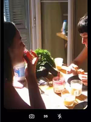 A woman is captured mid-bite, her mouth open as she enjoys a snack. She is indoors near a window with sunlight streaming in. A table is covered with food and drinks and another person's partially visible behind her.