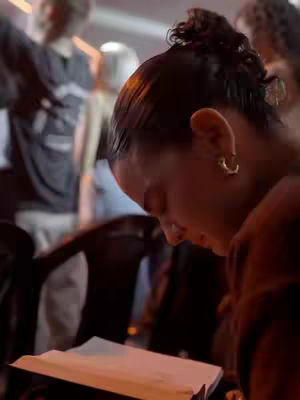 A woman is shown in a close-up shot, angled from the side, as she looks down, seemingly engaged in reading or writing on a paper. The background is blurred, but other people can be seen, suggesting a public setting. She is wearing a brown colored top, and has her hair up, with a golden hoop earrings, and a serious expression on her face.