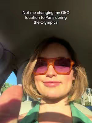 A selfie shows a woman in a car, wearing large orange sunglasses and a green top. The background is a bit blurred but suggests an outdoor setting with sunlight. Text overlayed on the image reads 'Not me changing my OkC location to Paris during the Olympics'.