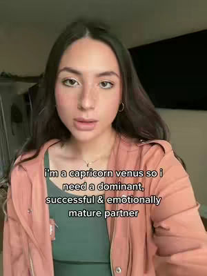 A woman with fair skin and long, dark hair is centered in the frame. She is wearing a green top and a light pink jacket. Text overlayed on the image reads: 'i'm a capricorn venus so i need a dominant, successful & emotionally mature partner'. The background is blurred, with neutral colors.