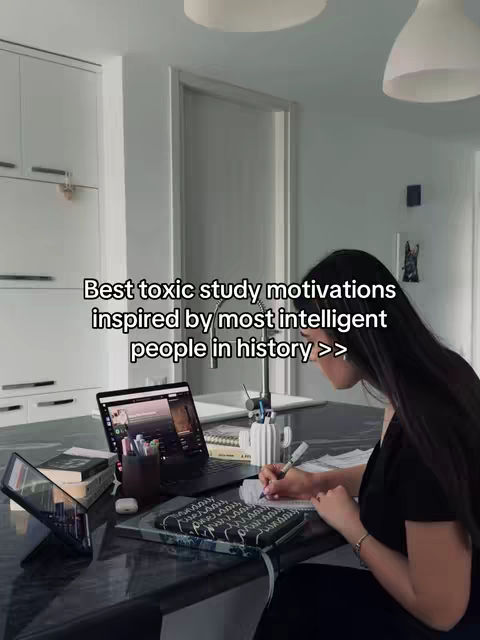 A person is working on a laptop and writing on a notebook in a kitchen. The scene is well-lit with overhead lights. A phone is resting angled to the left.