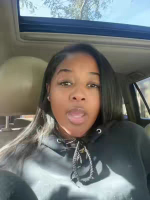 A woman is sitting in the driver's seat of a car, looking directly at the camera with her mouth slightly open. She has long, dark hair and is wearing a black hoodie. The background shows the interior of the car, including the sunroof and the headliner. The lighting appears soft and natural.