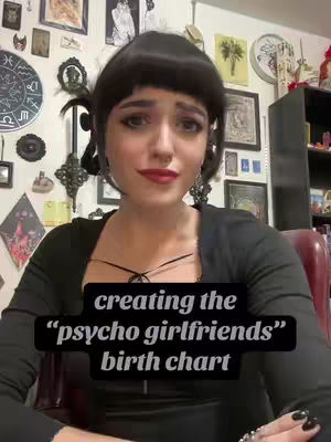 A woman with dark hair and makeup is centered in the frame, looking at the camera with a slightly concerned expression. She has a black top and is seated in front of a wall with various decorations and framed pictures. A text overlay in a black rectangle is across her chest.