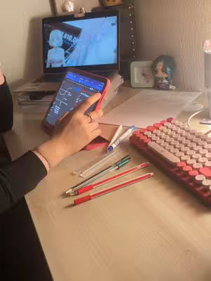 A person is sitting at a desk with a laptop and a tablet. The person is holding the tablet with one hand, and the screen of the tablet displays a chart of some sort. There are pens and a keyboard on the desk. A laptop is in the background with a cartoon on the screen.