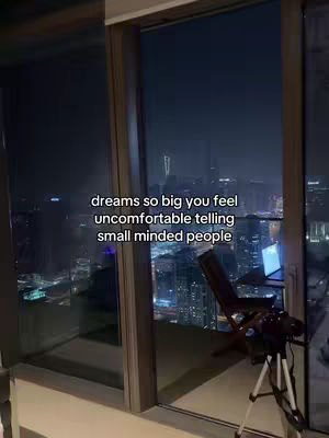 The image displays a nighttime cityscape viewed through a large window. The scene is dominated by the city lights, reflecting in the glass. In the foreground, there's an interior view with a wooden chair and a tripod with a camera set up, suggesting a recording space. Text, overlayed across the scene, reads: 'dreams so big you feel uncomfortable telling small minded people.' The overall composition conveys a sense of ambition and looking towards the future.
