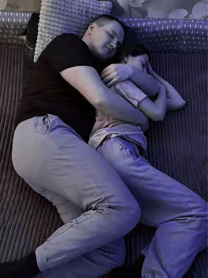 A color photo shows two people cuddling in bed. The person in the foreground is a man wearing a black t-shirt and gray sweatpants. He is embracing the person in the background, who seems to be a woman, wearing a light-colored shirt and pants, and covering their face with their hands, while they are both lying on the bed, with soft pillows.