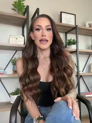 A woman is sitting in a chair and speaking to the camera with a bookshelf behind her. She is well-groomed with long, wavy brown hair and is wearing a black top and jeans. Several plants and framed pictures are on the shelves behind her.