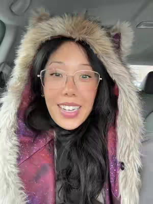 A woman with long black hair, wearing a furry hooded coat and glasses, smiles at the camera while sitting in a car.