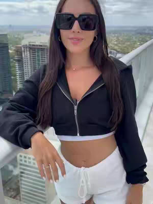 A woman wearing sunglasses and a black zip-up hoodie stands on a rooftop, with a cityscape in the background.