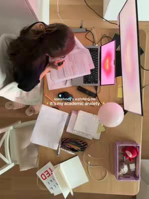 The cover image shows a top-down view of a desk, cluttered with various items, suggesting a workspace. A person, visible from the shoulders up, is leaning over the desk with dark hair partially obscuring their face. A laptop is open, displaying a screen with a pink and orange gradient. Several stacks of papers, pens, pencils, a mouse, a remote, and a pink balloon are scattered across the desk, creating a sense of focused activity. The lighting is soft and the overall tone appears to be casual and possibly intimate.
