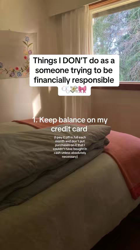 The video cover image shows a bedroom scene featuring a made bed with grey and pink bedding, a window on the right with sunlight streaming in, and a curtain that may be semi-sheer on the right side. A text overlay in a white rounded box with the words 'Things I DON'T do as a someone trying to be financially responsible' at the top occupies most of the top portion of the image. The text layer is above the main detail, which is a bulleted list item explaining the main advice or information about one point, 'Keep balance on my credit card', with an explanation below about how the creator manages their credit card in order to be financially responsible. To the left of the window, near the top, a small, framed, embroidered artwork featuring colorful floral designs adorns the wall.