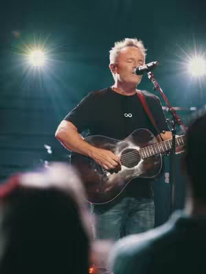 The cover image showcases a concert setting. A man with gray hair is centered, playing an acoustic guitar and singing into a microphone. Bright stage lights are positioned on either side, creating a spotlight effect. Several silhouettes of audience members are visible in the foreground. The color palette is centered around warm tones from the stage lighting, casting shadows and highlighting the performer.